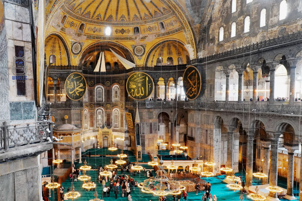 Wnętrze Hagi Sofii w Stambule – monumentalna kopuła ozdobiona złotymi mozaikami, islamskimi kaligrafiami i żyrandolami. Widok na przestrzeń modlitewną z dywanami oraz turystów zwiedzających to ikoniczne miejsce, które łączy chrześcijańskie i islamskie dziedzictwo kulturowe