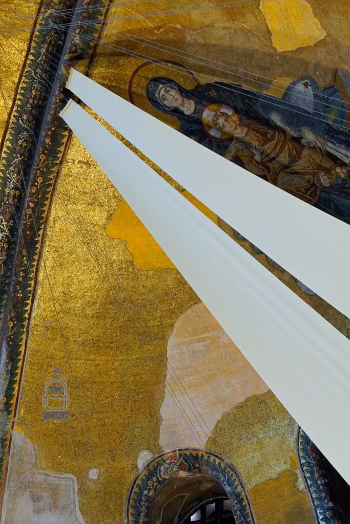 Fragment fresku z wizerunkiem Maryi i Dzieciątka w Hagia Sophia w Stambule, częściowo przysłonięty białymi tkaninami, widoczny na tle złotych mozaik.