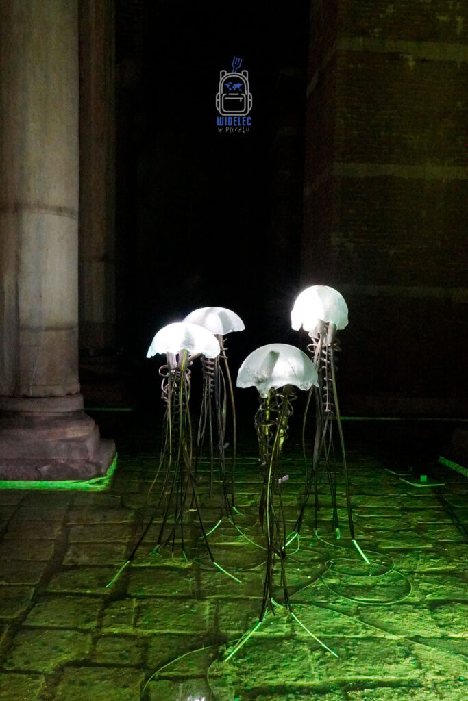 Artystyczna instalacja w kształcie meduz, podświetlona zielonym światłem, w podziemnych Cysternach Justyniana w Stambule, tworząca nowoczesny kontrast z historycznym otoczeniem marmurowych kolumn