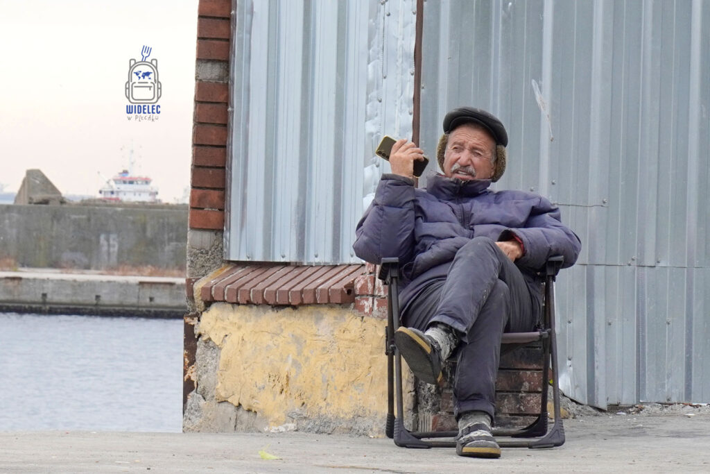 Zdjęcie ukazuje starszego mężczyznę siedzącego na krześle przy nabrzeżu w Stambule. Ubrany w ciepłą kurtkę i czapkę, trzyma telefon w ręku, spoglądając w dal z wyrazem zadumy. W tle widoczny jest fragment portu oraz przepływający statek. Obraz oddaje codzienność mieszkańców Stambułu i spokojne chwile nad brzegiem morza, gdzie tradycja spotyka się z nowoczesnością
