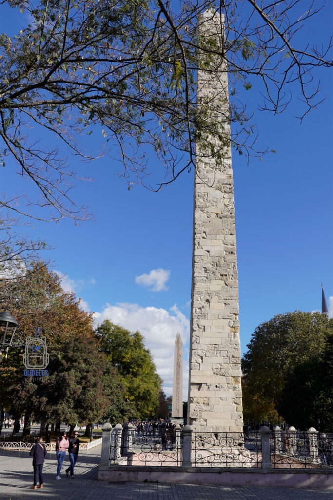 Obelisk Konstantyna na dawnym Hipodromie w Stambule, otoczony metalowym ogrodzeniem, na tle błękitnego nieba i zieleni drzew. W tle widoczny Egipski Obelisk Teodozjusza. Jedna z głównych atrakcji Placu Sultanahmet, łącząca historię Bizancjum z nowoczesnym Stambułem