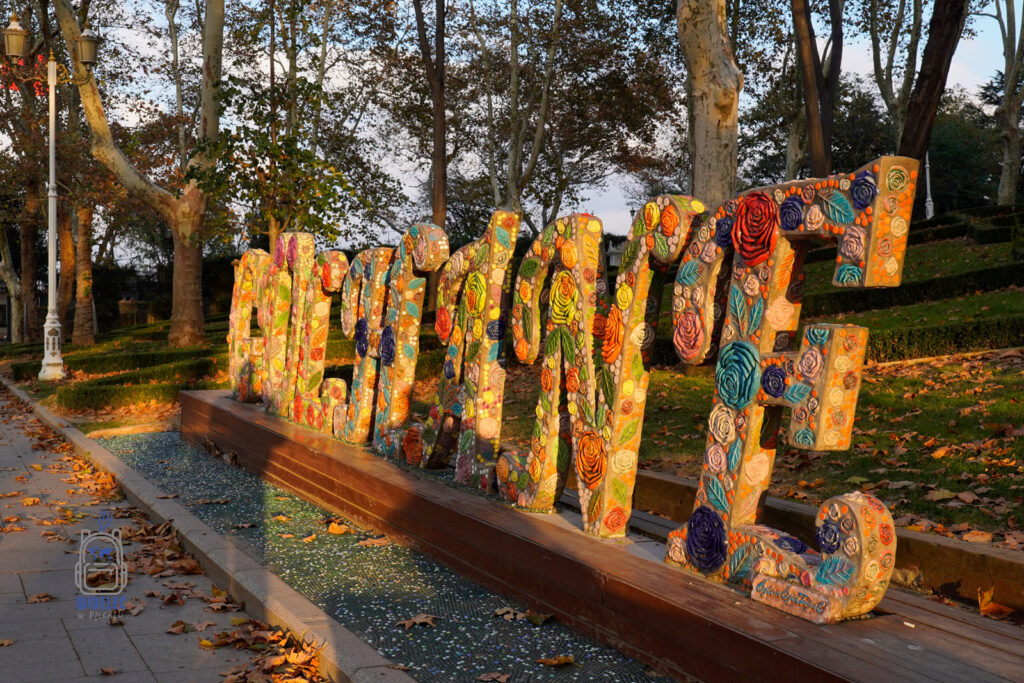 Kolorowa instalacja artystyczna z napisem "Gülhane" w parku Gülhane w Stambule, otoczona drzewami w jesiennej scenerii, ozdobiona wzorami kwiatów i mozaik.