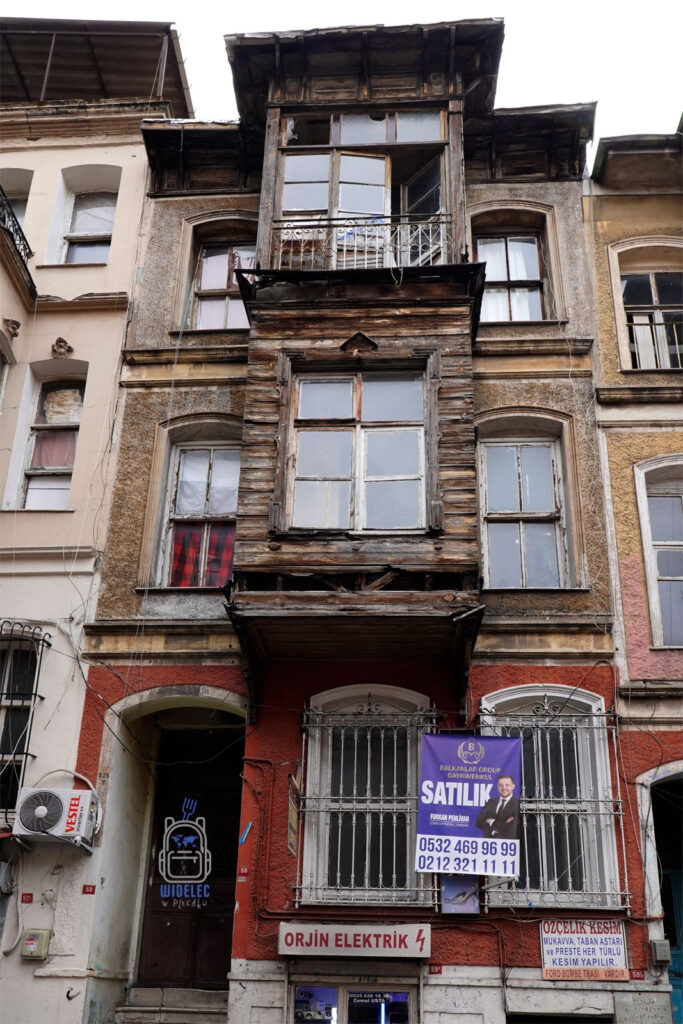 Zdjęcie przedstawia starą, drewnianą kamienicę w Stambule, położoną w jednej z historycznych dzielnic miasta. Budynek charakteryzuje się zniszczoną fasadą, drewnianymi elementami i typowymi dla tureckiej architektury wykuszami. Tablica 'Satılık' wskazuje, że nieruchomość jest na sprzedaż, co odzwierciedla zmieniającą się dynamikę lokalnego rynku nieruchomości. To miejsce pełne kontrastów – między przeszłością a nowoczesnością – oddaje klimat Stambułu, gdzie historia spotyka współczesność.