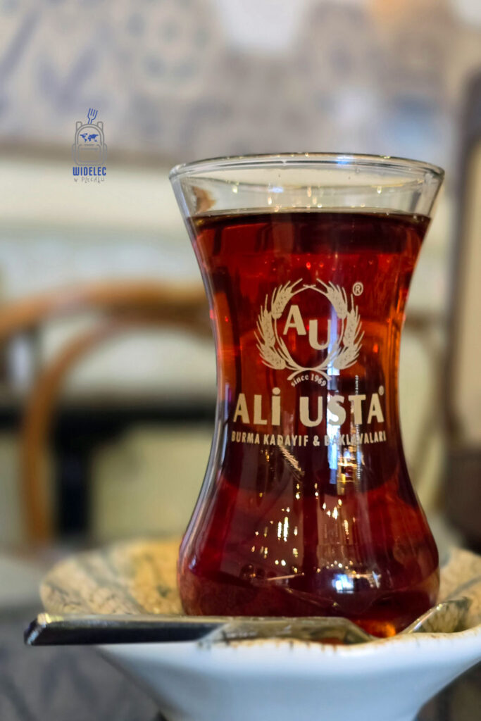 Tradycyjna turecka herbata podana w szklance w kształcie tulipana z logo "Ali Usta", stojąca na ozdobnym talerzyku w klimatycznej kawiarni w Stambule