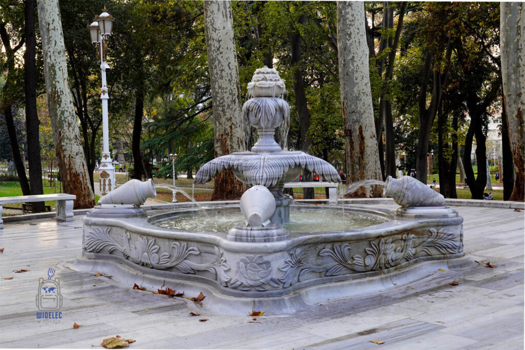 Elegancka marmurowa fontanna w parku Gülhane w Stambule, otoczona wysokimi drzewami i jesiennymi liśćmi, stanowiąca malowniczy element tego historycznego ogrodu.