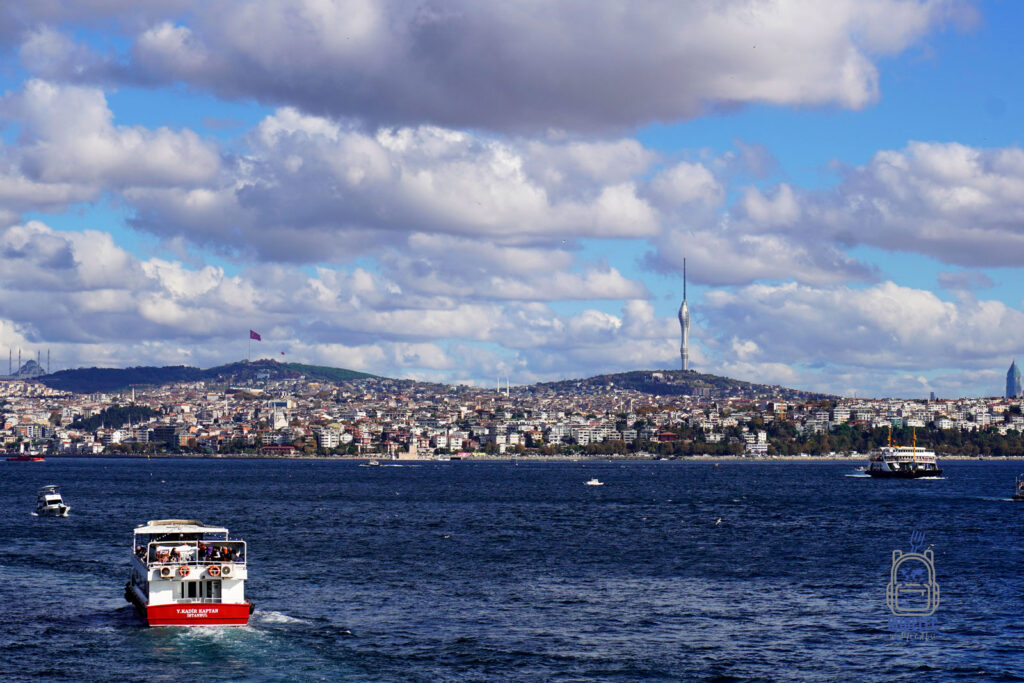 Zdjęcie przedstawia widok na Stambuł z perspektywy Bosforu. Na pierwszym planie znajduje się biało-czerwony prom pasażerski płynący przez lazurowe wody cieśniny, a w tle widać panoramę miasta z charakterystyczną wieżą telewizyjną Çamlıca Tower na wzgórzu. Obok niej widoczna jest turecka flaga powiewająca nad miastem. Krajobraz wypełniają gęsto zabudowane wzgórza, które oddają urok tętniącego życiem Stambułu. To miejsce, gdzie historia spotyka się z nowoczesnością, a piękno Bosforu zaprasza do odkrywania lokalnej kultury.