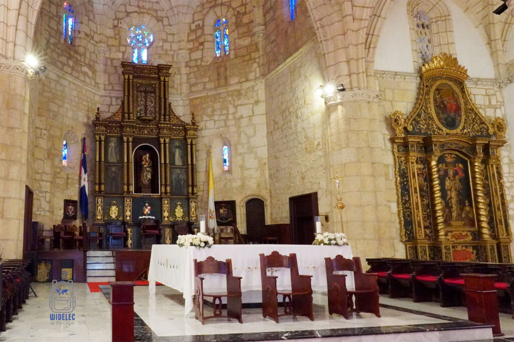 Zdjęcie przedstawia główny ołtarz Katedry Najświętszej Maryi Panny od Wcielenia w Santo Domingo. Ołtarz ozdobiony jest bogatymi, złotymi zdobieniami i obrazami religijnymi. Na środku widoczna jest figura Matki Bożej z Dzieciątkiem. Nad ołtarzem wyróżniają się kolorowe witraże, które dodają wnętrzu katedry barwnych akcentów. Po bokach ołtarza umieszczone są drewniane konfesjonały i krzesła z wysokimi oparciami. Całość otaczają kamienne ściany i sklepienia, dodając pomieszczeniu majestatycznego charakteru
Dominikana - z wschodu na zachód - z południa na północ