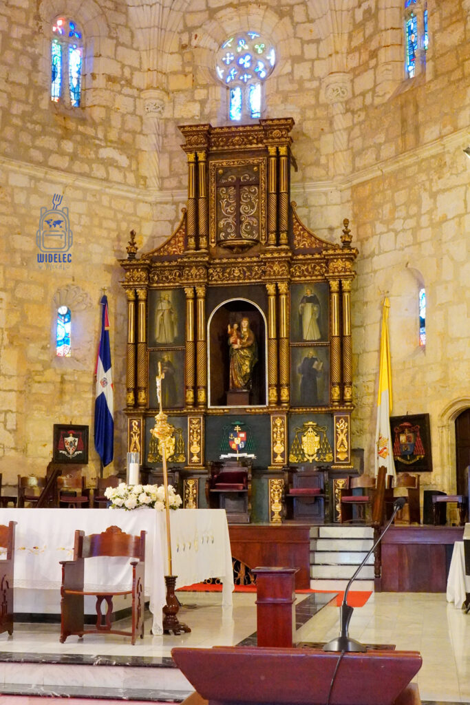 Zdjęcie ukazuje szczegółowy widok głównego ołtarza w Katedrze Najświętszej Maryi Panny od Wcielenia w Santo Domingo. Ołtarz zdobiony jest bogato rzeźbionymi, złotymi elementami oraz zawiera liczne obrazy religijne umieszczone w ramach. Centralne miejsce zajmuje figura Matki Bożej z Dzieciątkiem. Powyżej ołtarza widać kolorowe witraże, które wpuszczają do wnętrza katedry światło, tworząc efekt barwnych akcentów. Całość otoczona jest przez kamienny mur i łuki, nadając wnętrzu gotycki charakter. Na pierwszym planie widać białą ołtarzową sukienkę oraz drewniane krzesła używane podczas ceremonii
Dominikana - z wschodu na zachód - z południa na północ