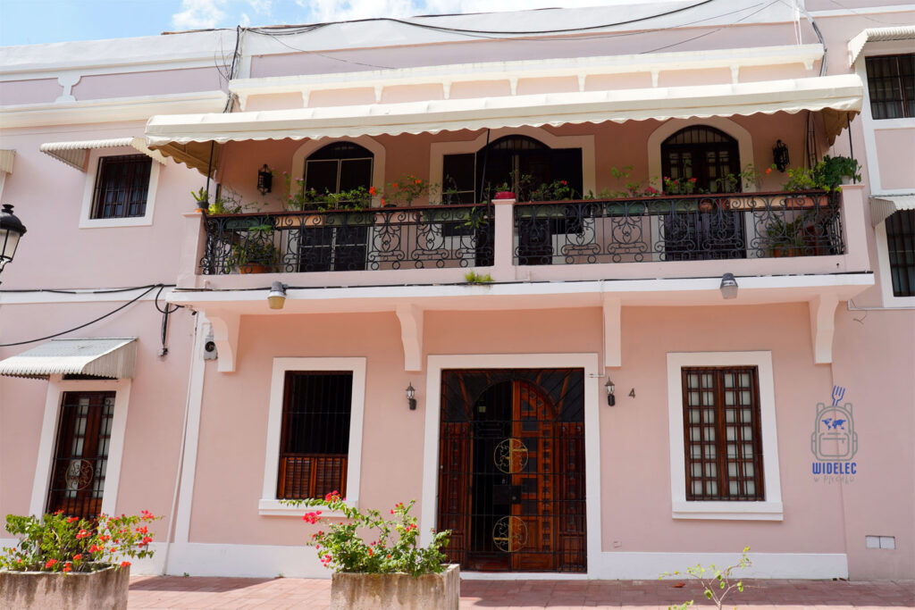 Zdjęcie przedstawia urokliwą, różową fasadę tradycyjnego domu w Santo Domingo, z dwoma poziomami, bogato zdobionymi balustradami i balkonami pełnymi kwiatów. Na parterze znajdują się drewniane drzwi z kutymi żelaznymi detalami oraz okno z drewnianymi okiennicami, a nad nimi szeroki, zacieniony balkon. Obok wejścia widoczne są kwitnące rośliny w donicach
Dominikana – Z wschodu na zachod - z południa na północ
