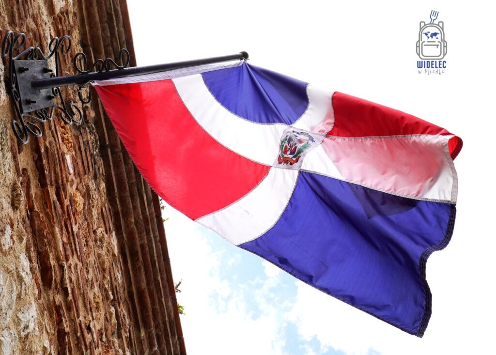 Flaga Republiki Dominikańskiej powiewająca na żelaznym maszcie przywiązana do starożytnej, ceglanej ściany
Dominikana - z wschodu na zachód - z południa na północ