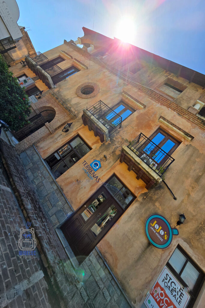 malownicza fasadę starego, okazałego budynku w dzielnicy kolonialnej Santo Domingo. Budynek charakteryzuje się pastelowo-żółtymi ścianami i zniszczonym tynkiem, co dodaje mu historycznego uroku. Widać także charakterystyczne, rustykalne detale architektoniczne, w tym drewniane drzwi i okiennice. Nad drzwiami umieszczono ozdobne logo firmy "Terrenas", a na balkonach rozmieszczone są doniczki z kwiatami. Kompozycja zdjęcia jest ujęta pod ostrym kątem, z słońcem świecącym bezpośrednio w obiektyw, co dodaje dynamiki i głębi scenie. Idealnie oddaje to klimat i estetykę kolonialnego Santo Domingo
Dominikana – Z wschodu na zachód - z południa na północ