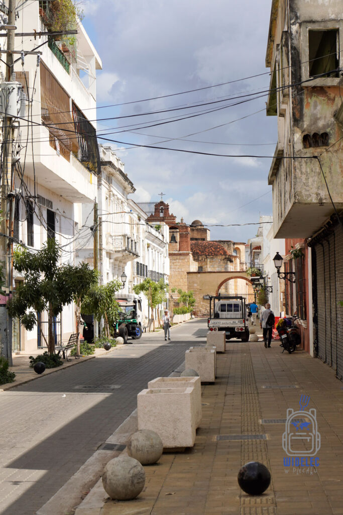 Zdjęcie ukazuje malowniczą ulicę w Santo Domingo, na której widać mieszankę architektury kolonialnej i nowoczesnej. Na pierwszym planie umieszczone są prostokątne, kamiennie donice, które nadają charakteru tej miejskiej przestrzeni. Białe, zdobione budynki z balkonami i latarniami kontrastują z bardziej zniszczonymi fasadami w tle, tworząc unikalny miejski krajobraz. W głębi widać starą, ceglaną bramę, która dodaje historycznego charakteru tej scenie. Na ulicy przechadzają się ludzie, co dodaje życia temu kadrze.
Dominikana – od wschodu do zachodu i z południa na północ