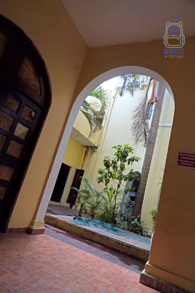 Zdjęcie ukazuje wnętrze otwartego patio w stylu kolonialnym w Santo Domingo, z widokiem przez łukowy przejazd. Scena zawiera bogatą roślinność, w tym palmy i niewielkie krzewy, które dodają zielonej świeżości kamiennemu otoczeniu. Terakotowe płytki na podłodze i ciemne, drewniane drzwi stanowią klasyczne elementy tej architektury
Dominikana – od wschodu do zachodu i z południa na północ