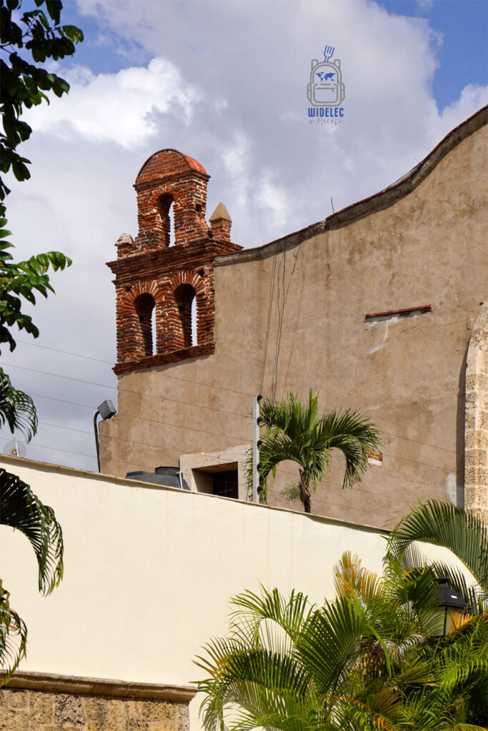 Zdjęcie prezentuje charakterystyczną dzwonnicę z czerwonej cegły, wznoszącą się nad białym murem w dzielnicy kolonialnej Santo Domingo. Dzwonnica ozdobiona jest klasycznymi arkadami i małymi, zaokrąglonymi szczegółami na szczycie, co dodaje jej tradycyjnego charakteru. W tle widać nieco zaniedbane, historyczne budynki, a pierwszy plan zdobi bujna zieleń tropikalnych roślin
Dominikana – Z wschodu na zachód - z południa na północ