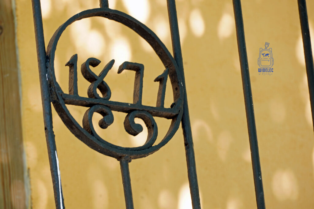 Zdjęcie prezentuje detal architektoniczny z numerem "1871" wykuszanym w ozdobnej, żelaznej kratce, stanowiącej element dekoracyjny budynku w Santo Domingo. Cyfry są elegancko uformowane, co nadaje elementowi unikatowego charakteru. Tło obrazu to jasna, żółtawa ściana, która kontrastuje z ciemnoniebieskim kolorem kraty, podkreślając jej detale i rok. Fotografia ta idealnie oddaje historyczny klimat miasta