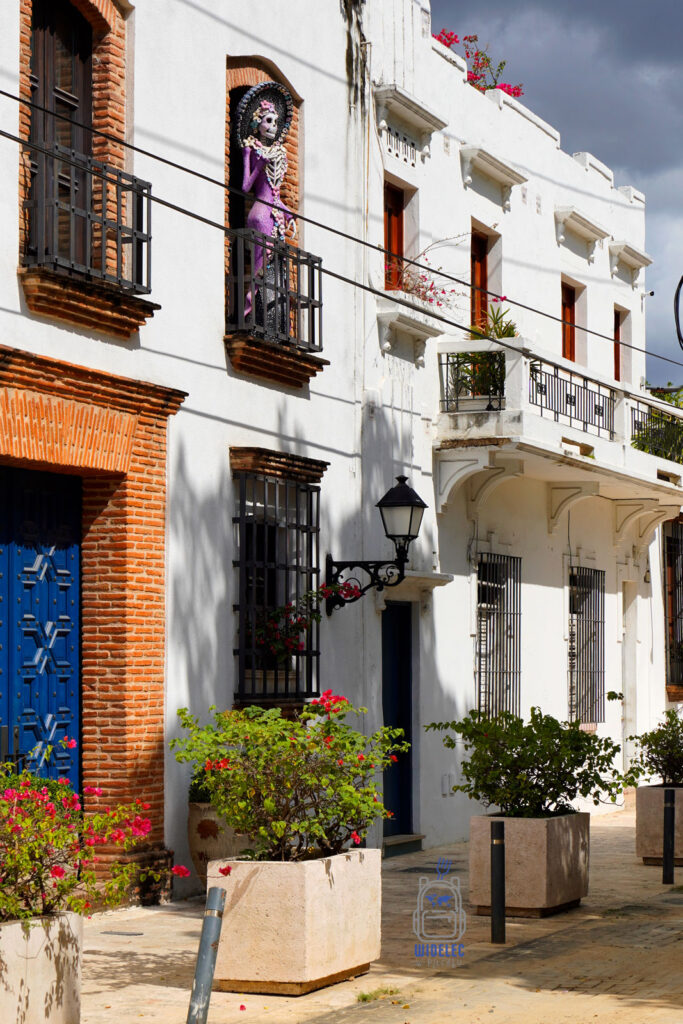 malownicza ulica w dzielnicy kolonialnej Santo Domingo, ukazująca klasyczne, białe fasady budynków zdobione ceglanymi detalami oraz kolorowymi drzwiami. Na jednym z balkonów  jest postać manekina przypominająca szkielet, ubrana w kolorowe stroje, co dodaje lokalnego, kulturowego charakteru scenie. Obfitość kwiatów i bujna zieleń roślin w donicach na chodniku dodatkowo podkreślają urok tego miejsca
Dominikana – Z wschodu na zachód - z południa na północ