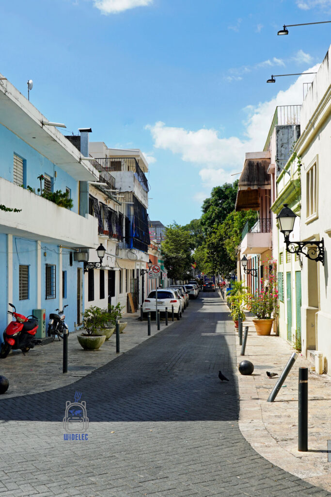 malownicza ulicę w dzielnicy kolonialnej Santo Domingo. Na zdjęciu są kolorowe, dwupiętrowe budynki z balkonami, przyozdobione donicami z kwiatami, które nadają ulicy żywy i przytulny charakter. Ulica jest wyłożona kamiennymi płytkami, a wzdłuż drogi zaparkowane są samochody i skutery. Na chodniku rosną zielone drzewa, co dodaje naturalnego cienia i uroku tej miejskiej przestrzeni. Ten widok jest idealny do przedstawienia na blogu podróżniczym "Widelec w Plecaku"
Dominikana – Z wschodu na zachód - z południa na północ
