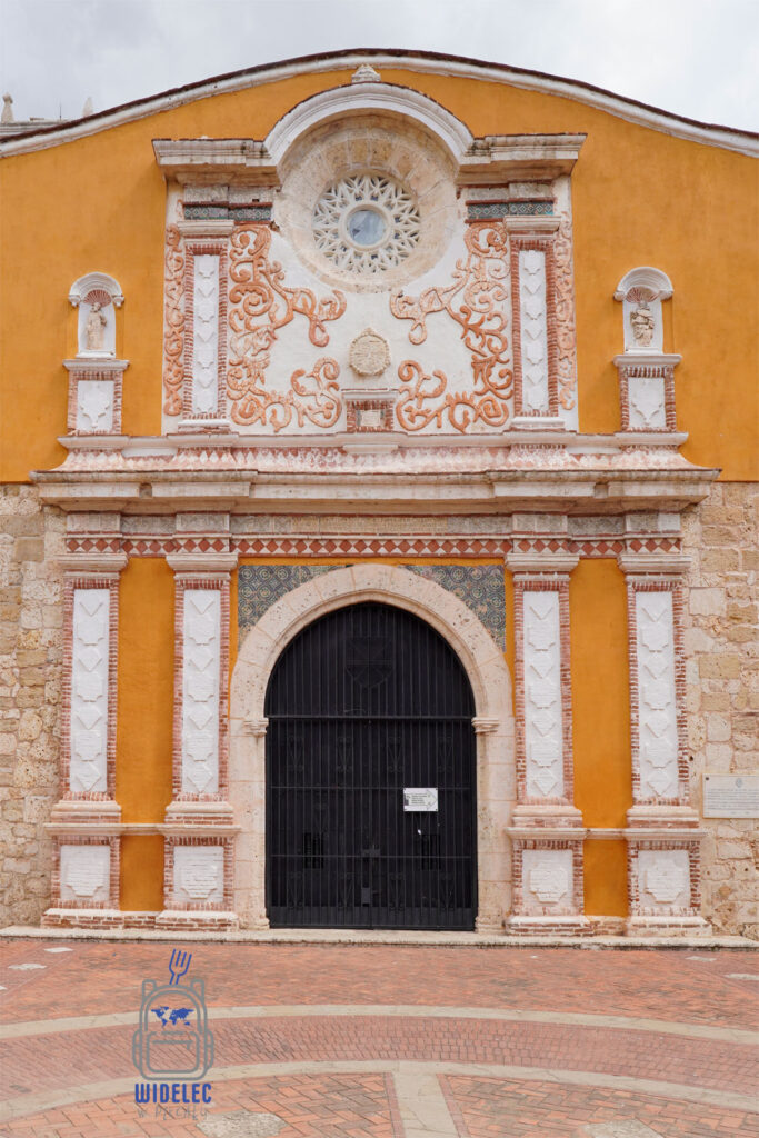 Zdjęcie przedstawia fasadę zabytkowego klasztoru dominikanów z 1532 roku w Santo Domingo. Budynek wyróżnia się bogato zdobioną, pomarańczową fasadą z wyrzeźbionymi detalami w kamieniu, które kontrastują z ceglano-białymi kolumnami i rozetą w centrum. Wielki, łukowaty portal prowadzi do wnętrza klasztoru, a całość stanowi doskonały przykład kolonialnej architektury
Dominikana – Z wschodu na zachód - z południa na północ