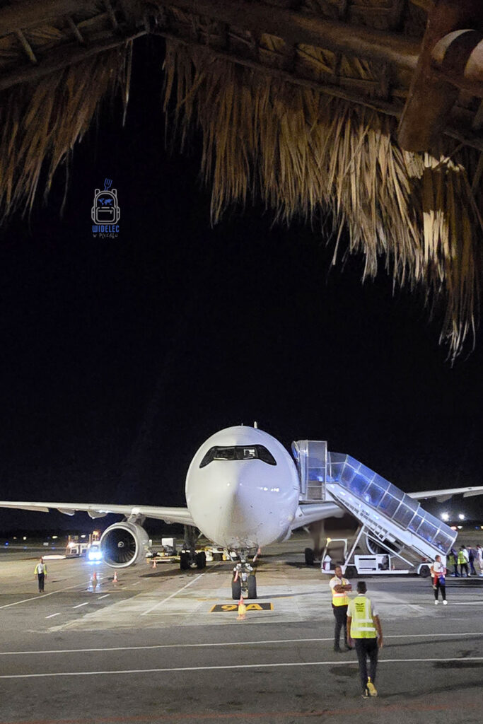 Samolot stojący na płycie lotniska w Dominikanie, nocą, przyświecają mu reflektory lotniska, które podkreślają jego wielkość i gotowość do dalszej podróży. W tle widać tradycyjny strzechowy dach, który dodaje lokalnego klimatu. Scena ta zachęca do podróżowania i odkrywania piękna Dominikany, 