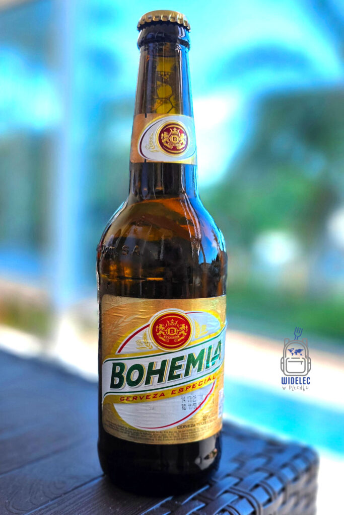 Piwo Bohemia w butelce stoi na tle błękitnego nieba i basenu, w samym sercu Dominikany. To popularne dominikańskie piwo typu pilsner jest cenione za swój złocisty kolor i bogaty, chmielowy smak. Idealne na ochłodę w gorących, tropikalnych warunkach
#widelecwpleacaku
#dominikana - z wschodu na zachód - z południa na północ