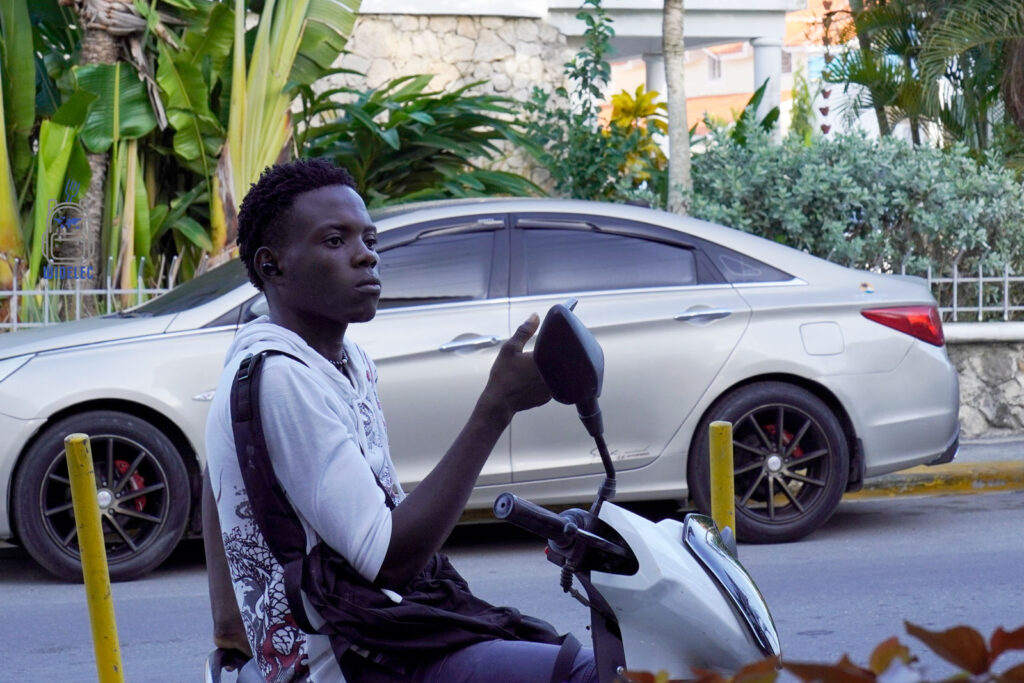 Młody mieszkaniec Dominikany, skupiony podczas jazdy na białym skuterze wśród miejskiej scenerii. Jego poważna postawa i stylowa, casualowa odzież, w tym biała koszulka i bordowa koszula na ramionach, uzupełniają dynamiczną atmosferę ulic Santo Domingo. Tło obrazu wypełniają bujne, zielone rośliny i eleganckie samochody, co dodaje zdjęciu autentyzmu miejskiego życia w Dominikanie
#widelecwpleacaku
#dominikana - z wschodu na zachód - z południa na północ