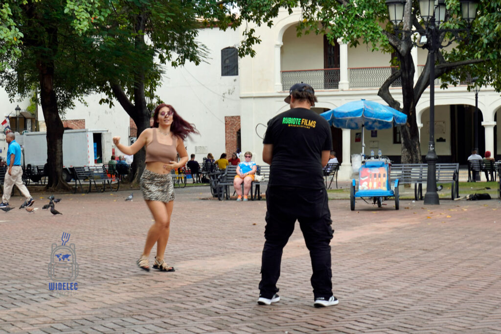 Młoda kobieta tańczy z pasją na jednym z placów Santo Domingo, podczas gdy mężczyzna filmuje ją kamerą. Tło obrazu wypełniają miejskie elementy: ławki, drzewa, i mijający przechodnie, co odzwierciedla energię i kulturową żywotność Dominikany. Na koszulce mężczyzny widoczne jest logo 'Rubiotte Films', co sugeruje profesjonalne nagrywanie filmów w publicznych miejscach
#widelecwpleacaku
#dominikana - z wschodu na zachód - z południa na północ