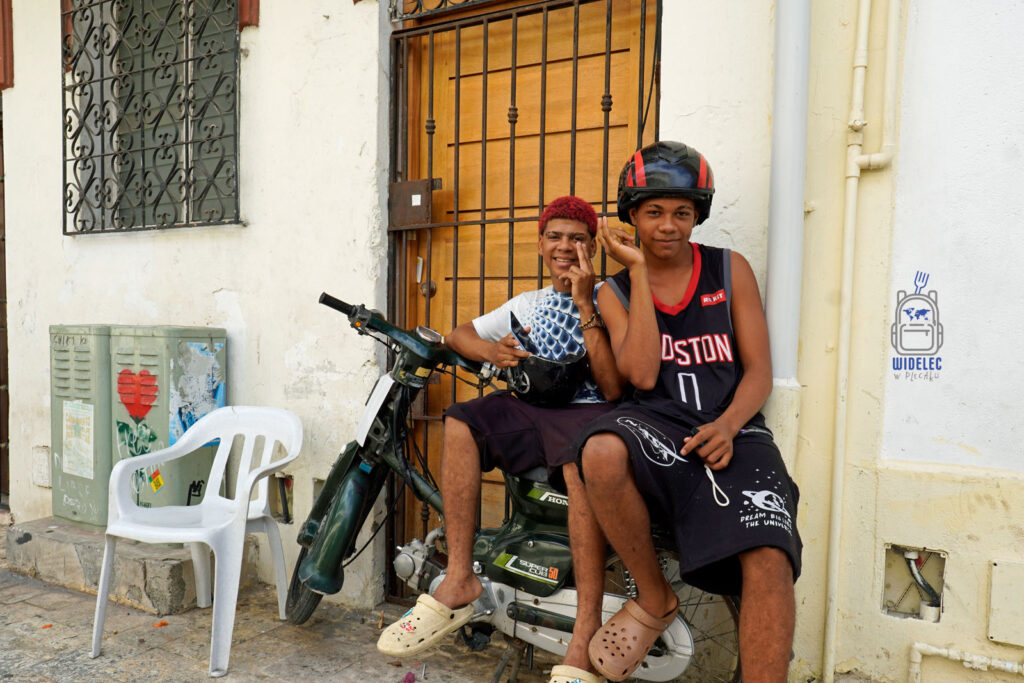 Dwóch młodych mieszkańców Dominikany siedzi na motorowerze przy brukowanej uliczce w Santo Domingo. Jeden z nich ma czerwone włosy i uśmiecha się do kamery, drugi, w czarnym bezrękawniku, ma na głowie kask. W tle widoczne są stare, białe ściany z kutymi kratami w oknach
#widelecwpleacaku
#dominikana - z wschodu na zachód - z południa na północ