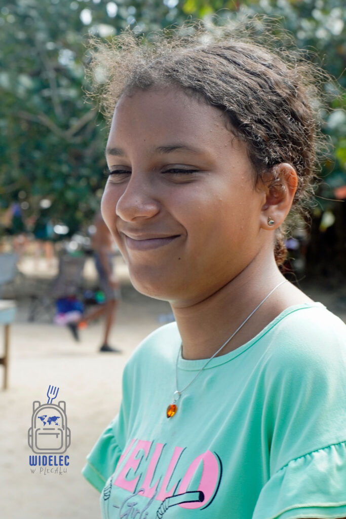 Urocza Viktoria, córka właściciela lokalnego hotelu w Dominikanie, uśmiecha się nieśmiało, stojąc na tle zatłoczonej plaży. Nosi jasnozieloną bluzkę z napisem 'HELLO' i delikatną srebrną naszyjnik