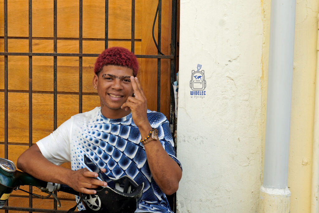 Młody mieszkaniec Dominikany z uśmiechem opiera się o motorower na ulicy Santo Domingo. Ma na sobie tradycyjną, kolorową koszulkę i czerwone włosy
#widelecwpleacaku
#dominikana - z wschodu na zachód - z południa na północ