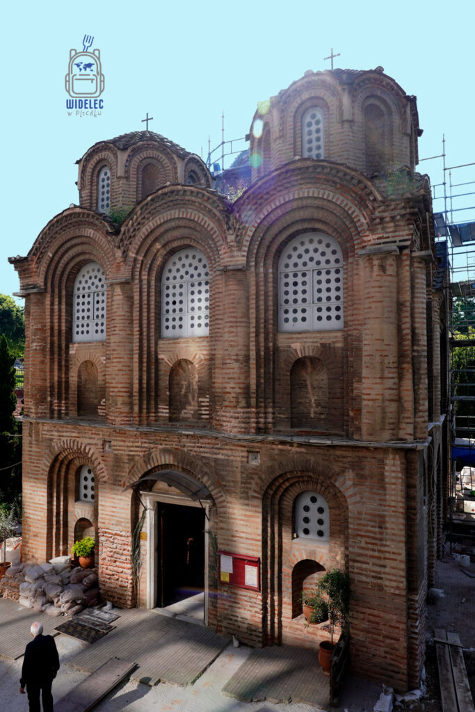 Bizantyjski kościół w Salonikach z cegły, z charakterystycznymi oknami i kopułami, otoczony rusztowaniami i roślinnością
