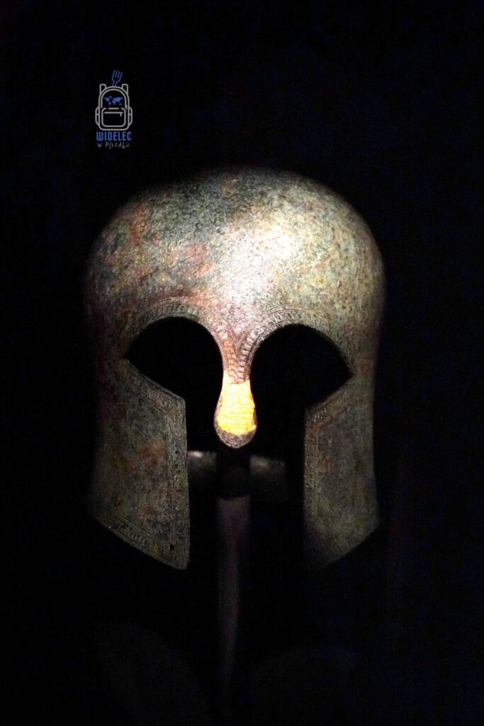 Starogrecki hełm hoplity z brązu, eksponowany w ciemności z subtelnym oświetleniem podkreślającym detale nad oczami i nos. Część ekspozycji w Muzeum Archeologicznym w Salonikach – pamiątka po czasach, gdy wojownicy piechoty greckiej budzili postrach w całym świecie antycznym.