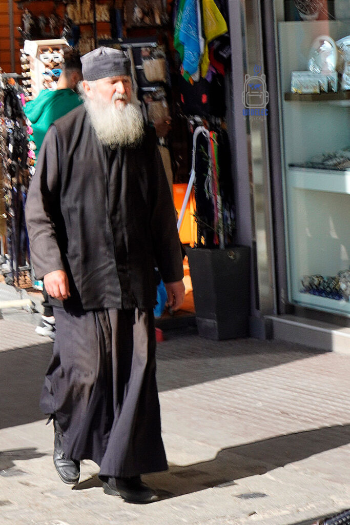 Prawosławny duchowny na tle sklepowej witryny w Salonikach – spotkanie tradycji i miejskiego zgiełku. Reportażowy kadr z Grecji na blogu Saloniki – Bogowie, Galeriusz i Gyros, widelecwplecaku.pl.