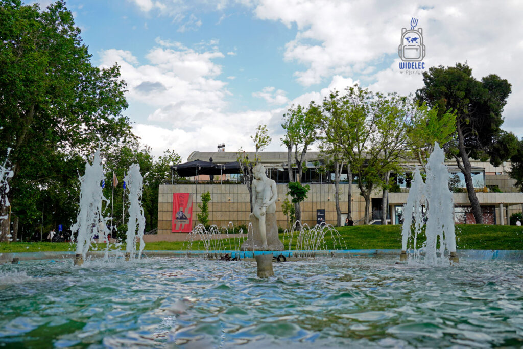 Fontanna z rzeźbą nagiego młodzieńca przed Muzeum Kultury Bizantyjskiej w Salonikach – nowoczesna architektura otoczona zielenią i wodnym detalem.