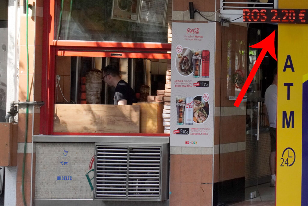 Uliczne okienko z gyrosem w Salonikach – klasyczny pionowy rożen z mięsem, oferta zestawu z pitą i Coca-Colą oraz widoczna cena 2,20 € za gyros na wynos.