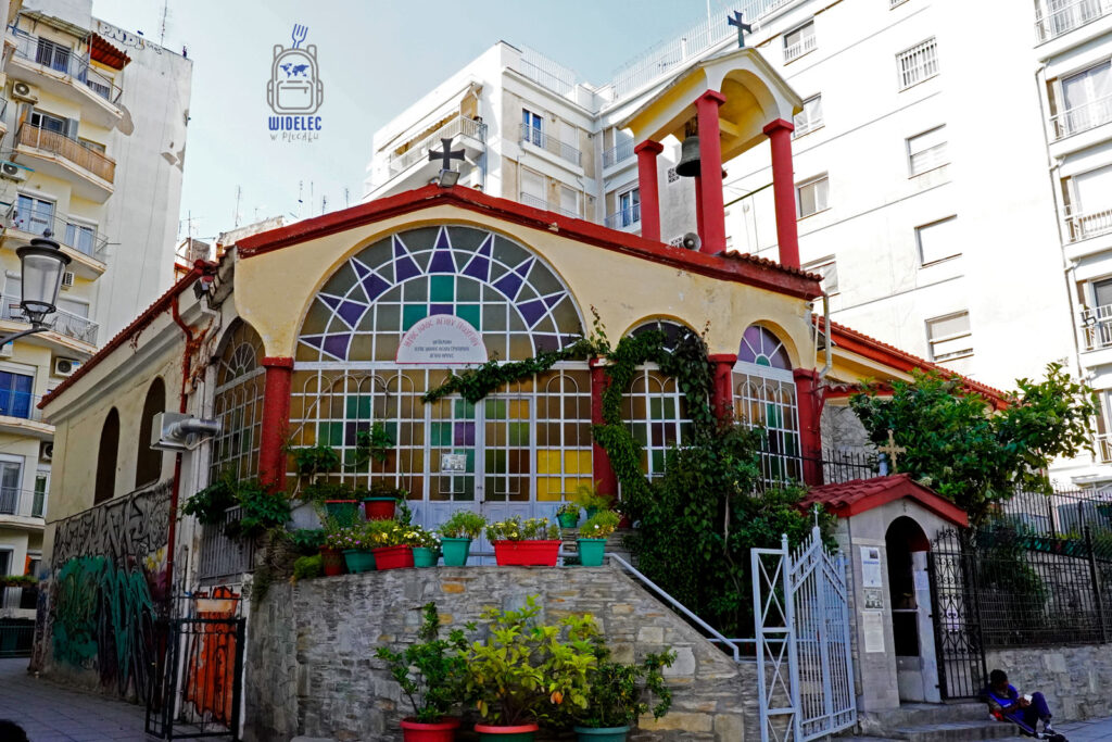 Kolorowa cerkiew z czerwonymi kolumnami i wielobarwnymi oknami, ukryta pomiędzy nowoczesnymi blokami w centrum Salonik – miejsce modlitwy w otoczeniu miejskiego zgiełku.