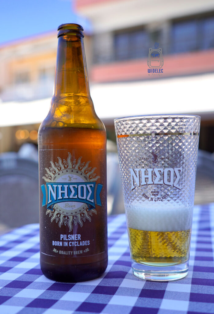 Butelka greckiego piwa Nisos i firmowy kufel na tle tawerny – lokalny pilsner z Cyklad, idealny do gyrosa i greckich smaków – Widelec w Plecaku.