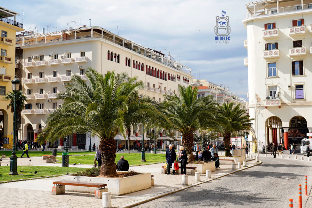 Plac Arystotelesa w Salonikach w słoneczny dzień – rzędy palm, spacerujący ludzie i kawiarniane stoliki otoczone elegancką architekturą z arkadami.