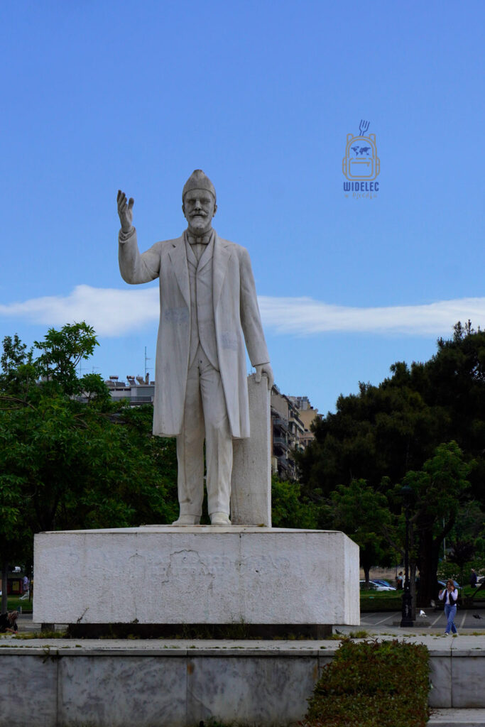 Pomnik Eleftheriosa Venizelosa w centrum Salonik – marmurowa statua przedstawiająca ojca współczesnej Grecji z uniesioną ręką, na tle miejskiej zieleni i zabudowy.