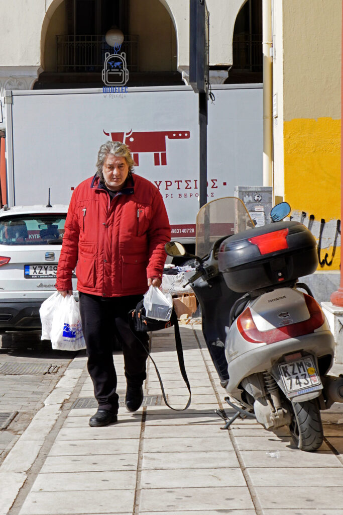 Starszy mężczyzna z zakupami na ulicy w Salonikach, w tle skuter i grecka codzienność – ujęcie miejskiego życia bez retuszu. Saloniki – Bogowie, Galeriusz i Gyros, blog Widelec w Plecaku.