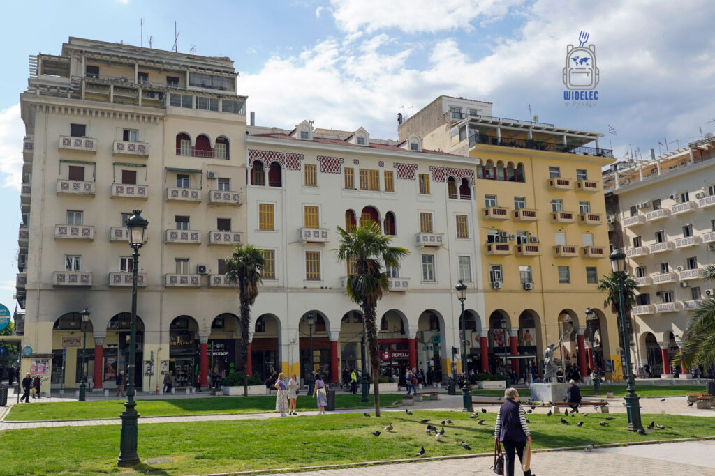 Plac Arystotelesa w Salonikach w pełnym słońcu – neoklasyczne budynki, palmy i mieszkańcy spacerujący po zielonym skwerze, w tle miejskie arkady i sklepy.