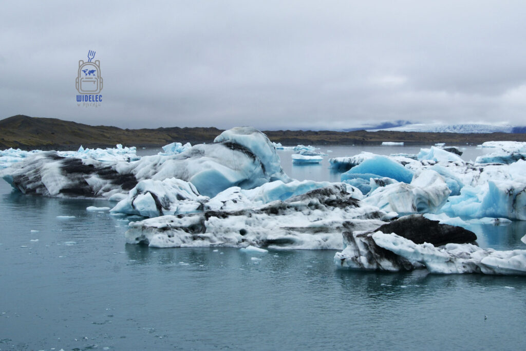 Islandia – lodowcowa laguna Jökulsárlón z turkusowymi i białymi górami lodowymi unoszącymi się na wodzie, w tle wzgórza i chmury; arktyczny pejzaż – widelecwplecaku.pl