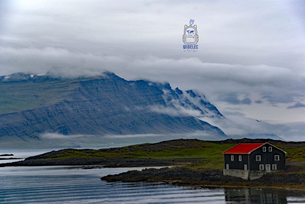 Islandia – samotny dom z czerwonym dachem stojący nad fiordem, w tle strome góry spowite mgłą i chmurami; surowy krajobraz północnej wyspy – widelecwplecaku.pl