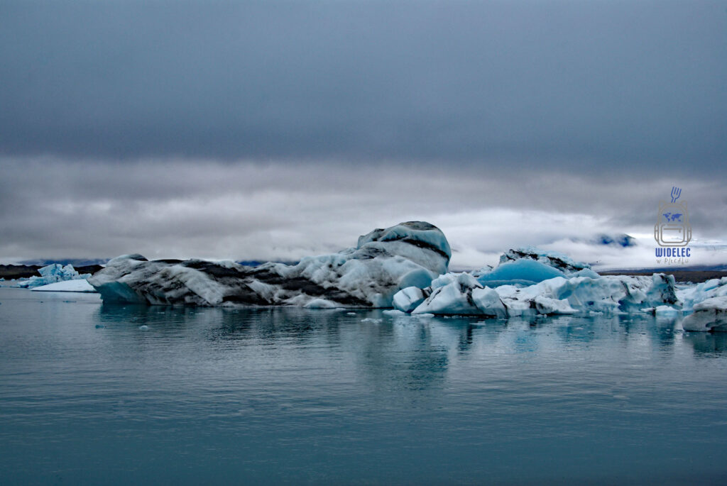Islandia – lodowe góry dryfujące w lagunie u podnóża Vatnajökull, turkusowe i białe bryły kontrastują z ciemnym niebem i chmurami; majestatyczny krajobraz – widelecwplecaku.pl