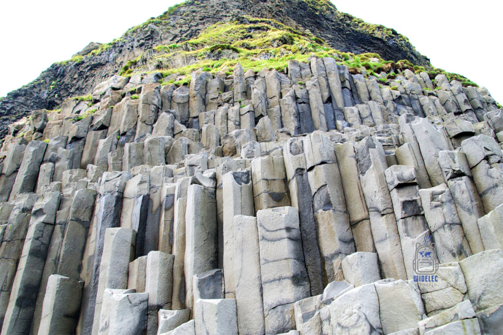 Naturalne bazaltowe kolumny na plaży Reynisfjara w Vik, Islandia – geometryczne formacje skalne tworzące imponującą ścianę kontrastującą z zielenią porastającą klif, widelecwplecaku.pl