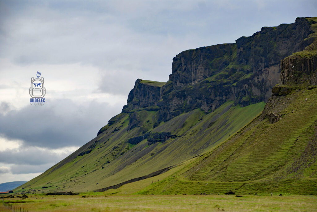 Islandia – strome klify i zielone zbocza gór porośnięte trawą, pod nimi rozległe łąki w surowym krajobrazie; monumentalna natura – widelecwplecaku.pl