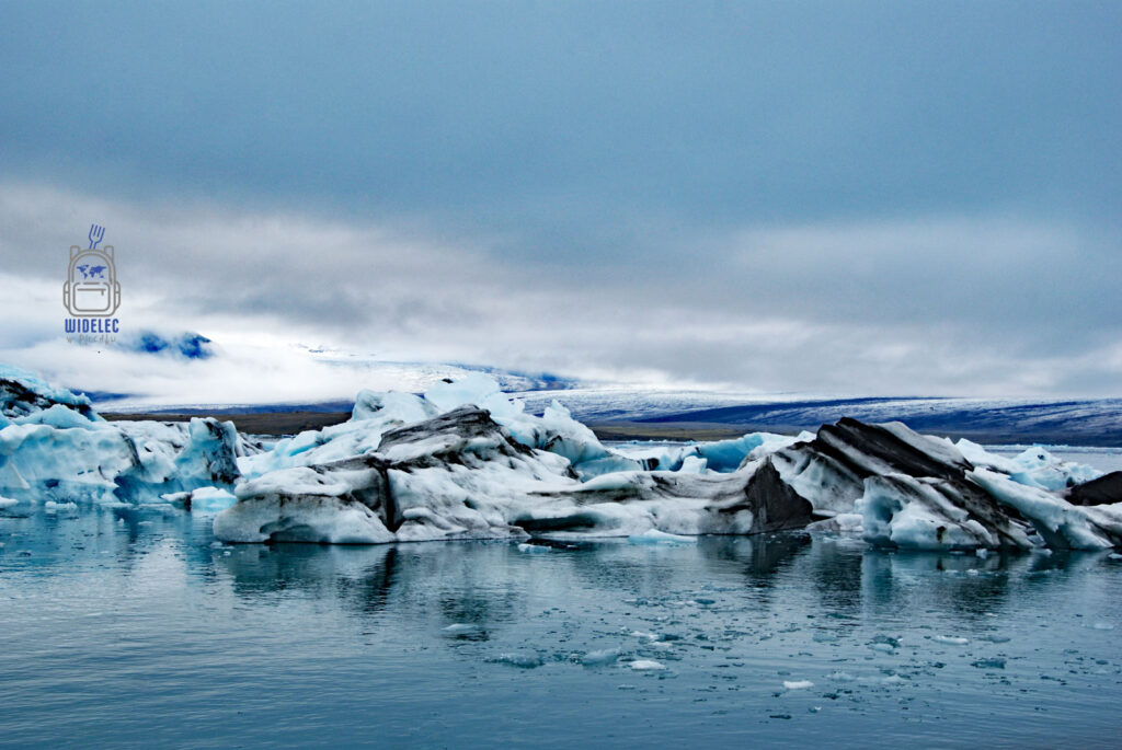 Islandia – lodowcowa laguna z dryfującymi turkusowo-białymi bryłami lodu, fragment lodowca Vatnajökull w tle pod chmurami; surowe piękno natury – widelecwplecaku.pl
