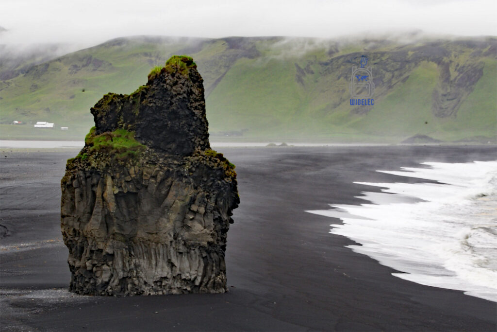 Islandia – czarna plaża Reynisfjara z bazaltową skałą wynurzającą się z piasku, kontrastującą z zielonymi wzgórzami i białą pianą fal; magiczny krajobraz południowego wybrzeża – widelecwplecaku.pl