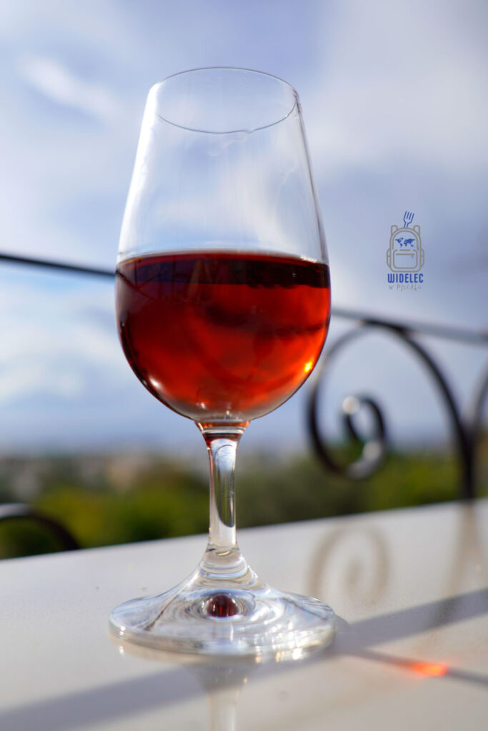 Kieliszek słodkiej commandarii – tradycyjnego cypryjskiego wina z podsuszanych winogron, o głębokim bursztynowym kolorze i nutach miodu oraz suszonych owoców, degustowany na tarasie z widokiem na wyspę Cypr