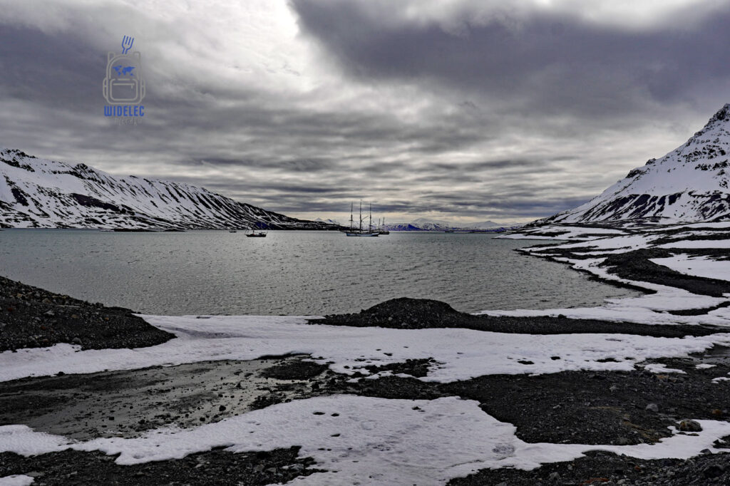 fiord z żaglowcem i mniejszą łodzią wokół ośnieżone szczyty pod przykrywką ciężkiego granatowego nieba. Svalbard, Arktyka