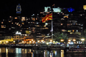Nocna panorama Ochrydy w Macedonii Północnej, widziana od strony Jeziora Ochrydzkiego. Na pierwszym planie powiewa duża, czerwono-żółta flaga Macedonii Północnej, a w tle na wzgórzu wznosi się monumentalna twierdza cara Samuela, efektownie podświetlona na biało i niebiesko. Poniżej twierdzy rozciąga się zabytkowa zabudowa miasta, której okna i uliczki rozświetlają liczne ciepłe światła. Wzdłuż nabrzeża widoczne są restauracje, spacerujący ludzie, latarnie oraz zacumowane łodzie. Kolorowe refleksy miejskich świateł odbijają się w ciemnej tafli jeziora. Fotografia oddaje tętniącą życiem, wakacyjną atmosferę Ochrydy po zmroku, łącząc historyczny charakter miasta z urokiem nocnego wypoczynku nad wodą.